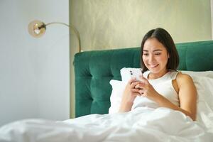 hermosa sonriente asiático mujer en cama, leyendo mensaje en teléfono inteligente y reír, disfrutando perezoso día en dormitorio, utilizando móvil teléfono foto