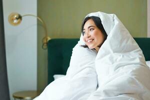 Cozy mornings. Happy asian girl feels warm in her bed, covers herself with cozy duvet in her bedroom, a comfortable stay in hotel room photo