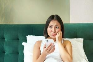 Portrait of sad frowning korean girl, lying in bed and looking at smartphone with upset, disappointed face expression, using mobile phone, complaining photo