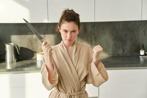 Close up of serious and confident brunette woman holds knife and clenches fist, standing in the kitchen, looking self-assured, posing in bathrobe photo