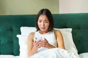 Bedroom picture of asian woman lying in bed, looking scared and shocked at smartphone screen, reading message with concerned face photo