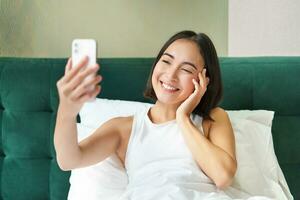 Beautiful asian girl lying in bed, making morning selfie, taking picture on smartphone in bedroom, smiling happily photo