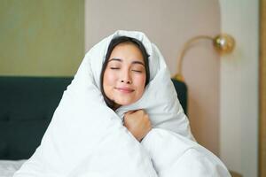 Smiling cute asian woman hugs her warm and cozy duvet, feels warm, makes cocoon from blanket, sits in her bedroom in early morning photo
