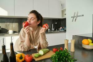 Portrait of beautiful young woman smelling tomatoes, cooking salad from fresh vegetables bought in market, preparing food for family, lead active and healthy lifestyle, standing in kitchen photo