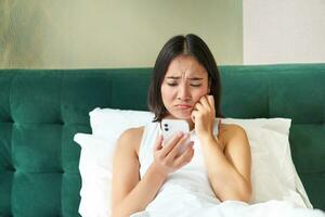 retrato de triste ceñudo coreano chica, acostado en cama y mirando a teléfono inteligente con decepcionado, decepcionado cara expresión, utilizando móvil teléfono, quejumbroso foto