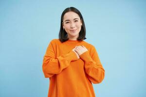 belleza y protección de la piel. cerca arriba retrato de contento sonriente japonés mujer, toques su claro, brillante piel, natural sano rostro, en pie terminado azul antecedentes foto