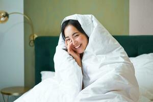 Beautiful asian woman sitting on bed, covered in blankets and duvet, laughing and smiling, enjoying weekend in bedroom photo