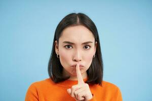 Close up of angry brunette asian girl, shush, touches lips with finger, tells to be quiet, stands over blue background photo