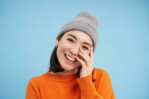 belleza y protección de la piel. cerca arriba retrato de contento sonriente japonés mujer, toques su claro, brillante piel, natural sano rostro, en pie terminado azul antecedentes foto