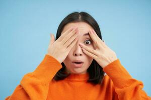 Close up portrait of korean girl waits for surprise, shuts her eyes and peeks through fingers with excited smile, stands over blue background photo