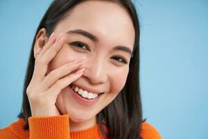 belleza y protección de la piel. cerca arriba retrato de contento sonriente japonés mujer, toques su claro, brillante piel, natural sano rostro, en pie terminado azul antecedentes foto