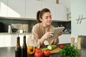 Portrait of woman checking recipe notes in notebook, standing in kitchen with vegetables, cooking food, preparing delicious salad from tomatoes, parsley and olive oil photo