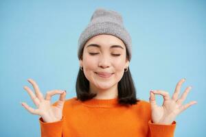 Sounds mind in healthy body. Smiling calm and relaxed asian girl in beanie, shows zen, relaxation gesture, meditating, standing over blue background photo