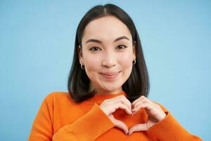 Close up portrait of lovely asian woman, shows heart sign and smiles with care and tenderness, stands over blue background photo