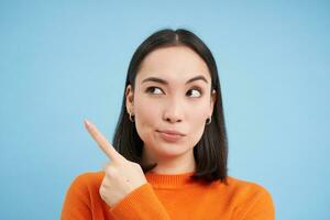 Close up of skeptical asian woman, looks and points left with suspicious face, stands over blue background photo