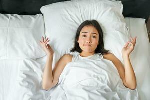 Portrait of asian girl lying in her bed and shrugs, looks puzzled, overthinking before going to sleep, looking complicated at camera photo