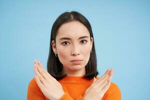 Close up portrait of serious asian woman, shows stops sign, disapproves something, refuses or rejects, stands over blue background photo