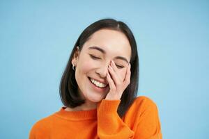 cerca arriba de hermoso morena mujer, riendo y sonriente, toques su rostro, expresa puro alegría, soportes terminado azul antecedentes foto