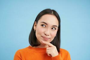 Close up of thinking asian girl, looks pleased and touches chin thoughtful, stands over blue background photo