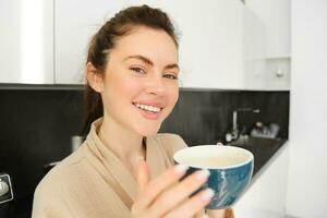 retrato de guapo joven mujer comenzando su día con taza de café, en pie en el cocina y Bebiendo capuchino desde grande taza, disfrutando favorito bebida en el Mañana foto