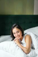 Vertical shot of smiling korean woman lying on pillow in her bed, spending morning in bedroom, waking up from sleep, looking happy photo