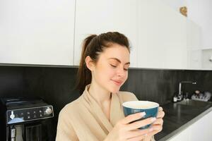 retrato de guapo joven mujer comenzando su día con taza de café, en pie en el cocina y Bebiendo capuchino desde grande taza, disfrutando favorito bebida en el Mañana foto