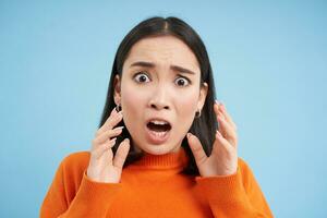 Close up of shocked asian woman in panic, anxious, looks terrified, stands over blue studio background photo