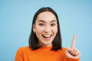 Close up portrait of enthusiastic asian woman, points up and laughs, smiles and looks happy, stands over blue studio background photo