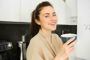 retrato de guapo joven mujer comenzando su día con taza de café, en pie en el cocina y Bebiendo capuchino desde grande taza, disfrutando favorito bebida en el Mañana foto