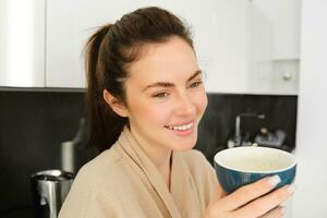 retrato de guapo joven mujer comenzando su día con taza de café, en pie en el cocina y Bebiendo capuchino desde grande taza, disfrutando favorito bebida en el Mañana foto