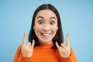 Close up of happy korean woman, shows rock n roll, heavy metal gesture, having fun, standing over blue studio background photo