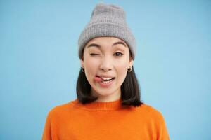 elegante asiático mujer en gorro, muestra lengua y guiños a tú, mira despreocupado, soportes en naranja camisa de entrenamiento terminado azul antecedentes foto