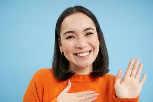 cerca arriba de simpático sonriente japonés mujer, levanta uno brazo, introducir sí misma, saluda, soportes terminado azul antecedentes foto