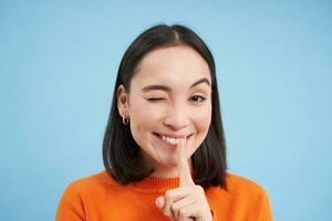 Close up of smiling asian girl, winks at you, tells secret, holds finger on lips, stands over blue studio background photo