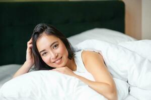 Portrait of young korean woman smiling, lying in bed on pillow, posing in her bedroom, looking relaxed and feeling comfortable photo