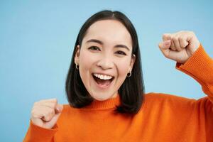 cerca arriba de alegre japonés mujer celebra, levanta manos arriba y risas, gana y triunfos, soportes terminado azul antecedentes foto