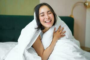 hermosa asiático mujer sentado en cama, cubierto con blanco funda Nordica, sonriente, disfrutando contento fin de semana mañana, riendo a cámara foto