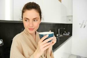 retrato de guapo joven mujer comenzando su día con taza de café, en pie en el cocina y Bebiendo capuchino desde grande taza, disfrutando favorito bebida en el Mañana foto