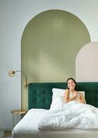 Vertical shot of beautiful asian girl lying in bedroom with sophisticated interrior, smiling and looking happy in morning, spending time in bed photo