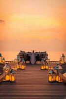 Amazing romantic dinner on the beach on wooden deck candles under sunset sky. Romance and love, luxury destination dinning, exotic table setup with sea view. Honeymoon proposal design photo