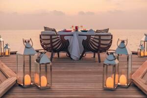Amazing romantic dinner on the beach on wooden deck candles under sunset sky. Romance and love, luxury destination dinning, exotic table setup with sea view. Honeymoon proposal design photo