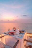 Amazing romantic dinner on the beach on wooden deck candles under sunset sky. Romance and love, luxury destination dinning, exotic table setup with sea view. Honeymoon proposal design photo