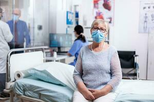 Senior woman in hospital room with face mask for portection against covid19 waiting medical test results. Global health crisis, medical system during pandemic, sick elderly patient. photo