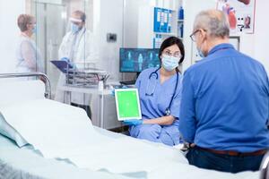 Nurse with face mask for protection against covid 19 holding tablet with green mockup while consulting senior man. Medical examination for infections, disease and diagnosis. photo