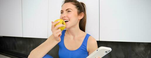 Image of stylish sportswoman, young fitness instructor sitting in kitchen and eating an apple, holding smartphone, using social media app on mobile phone photo