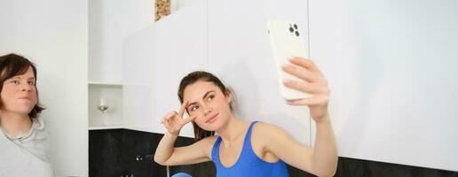 Portrait of smiling, beautiful fitness instructor, posing for fitness blog, selfie on smartphone, sitting in kitchen, looking happy photo