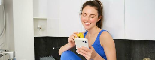 Healthy lifestyle and technology. Young beautiful girl, fitness instructor, looking at smartphone, reading on mobile phone and eating an apple, smiling happily photo