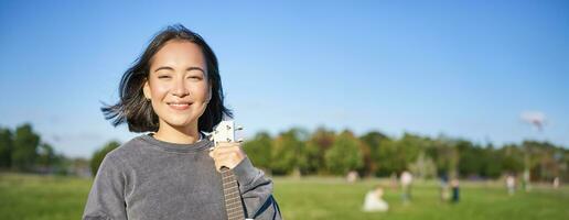 vertical Disparo de joven mujer muestra su ukelele, sostiene teléfono inteligente en mano y mira a cámara foto