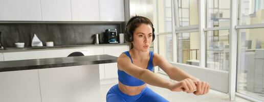 Image of young woman doing aerobics workout form home, listening music in wireless headphones, making squats exercises, fitness in living room photo