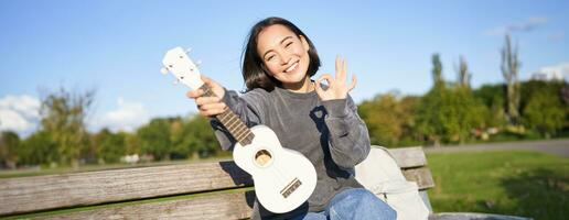 Cute smiling girl shows ok sign and her new ukulele, sits on bench in park, recommends musical instrument photo